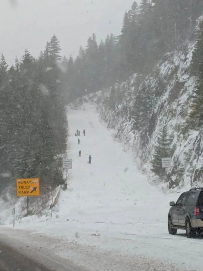 People walking up a snow-covered runaway truck ramp, a dangerous and unnecessary activity, with a vehicle in the foreground.