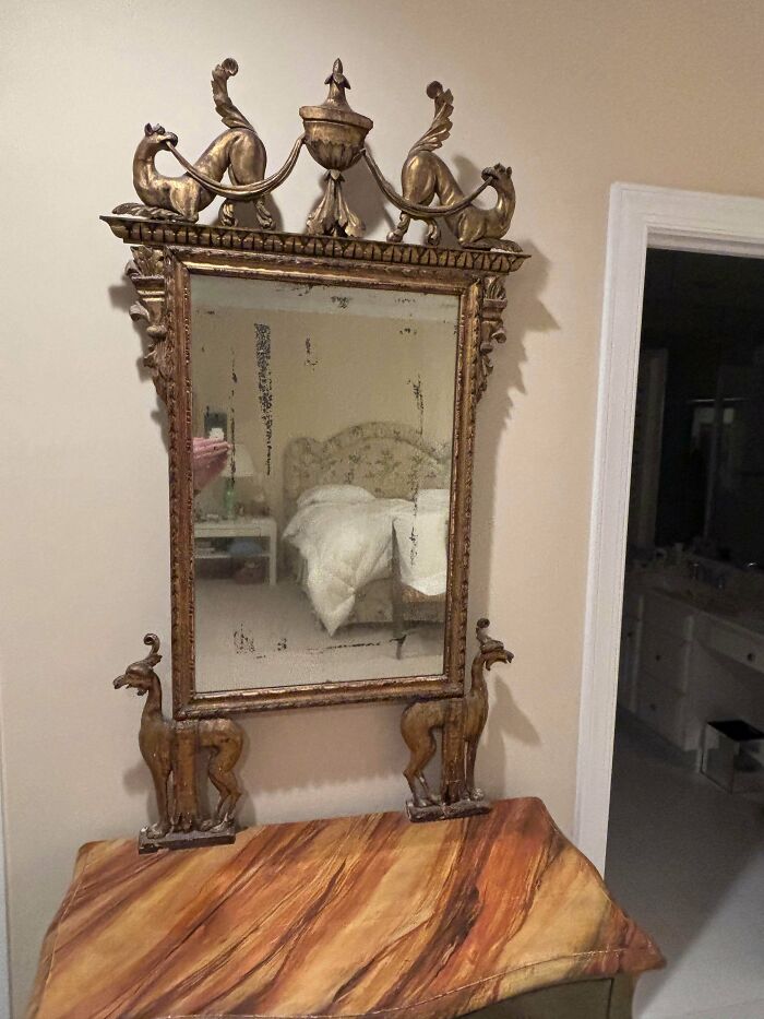 A remarkable antique gilded mirror with decorative animal figures at the top and base, reflecting a bedroom.