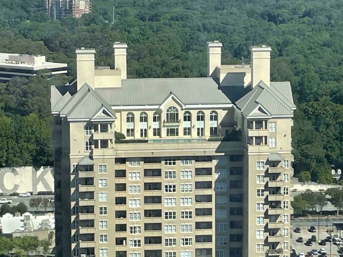 High-rise building with mansion-like elements on the top floors, featuring ornate roofs and windows, against a backdrop of trees.