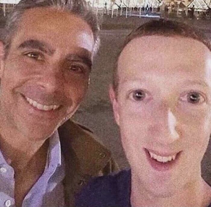 Mark Zuckerberg with a wide, unnatural smile next to another man, both having a bad hair day.