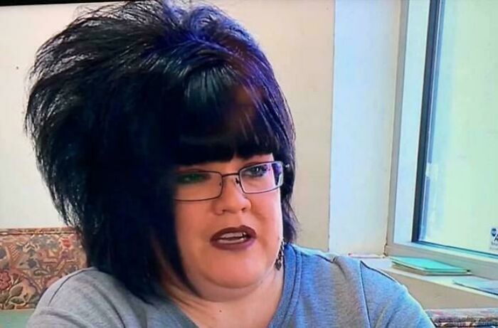 A woman with an extremely voluminous, dark, and purple-streaked hairstyle, wearing glasses, having a bad hair day.