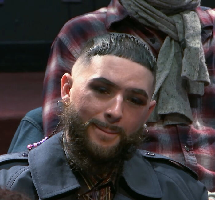A man with a precise yet unusual haircut and beard, wearing makeup, showcasing a bad hair day.