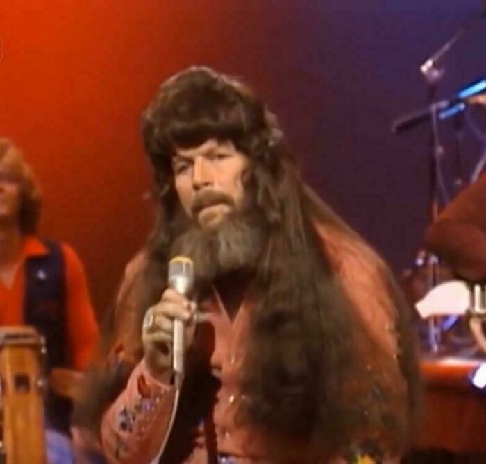 A man on stage with an extremely long, curly wig and a beard, making for a bad hair day.