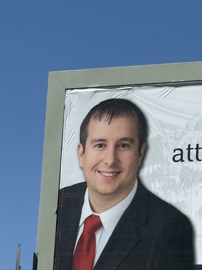A billboard of a man with a ridiculous bad hair day, wearing a suit and red tie, against a blue sky.
