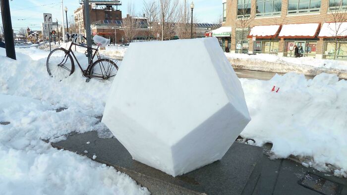 A large, interesting, geometric snow sculpture on a sidewalk next to a bicycle and piles of snow.