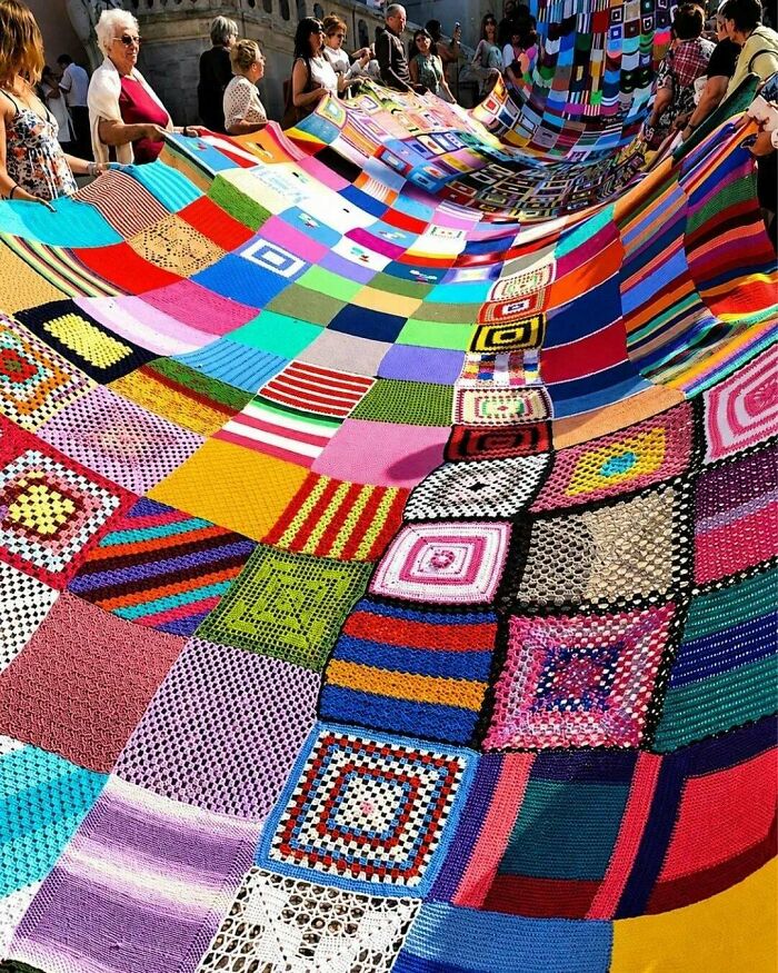 People holding a giant, colorful knitted blanket, an interesting photo of a community project.