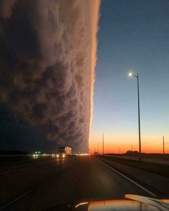 An interesting dramatic wall of clouds looms over a highway at dusk, with the sky showing a gradient of light to dark.