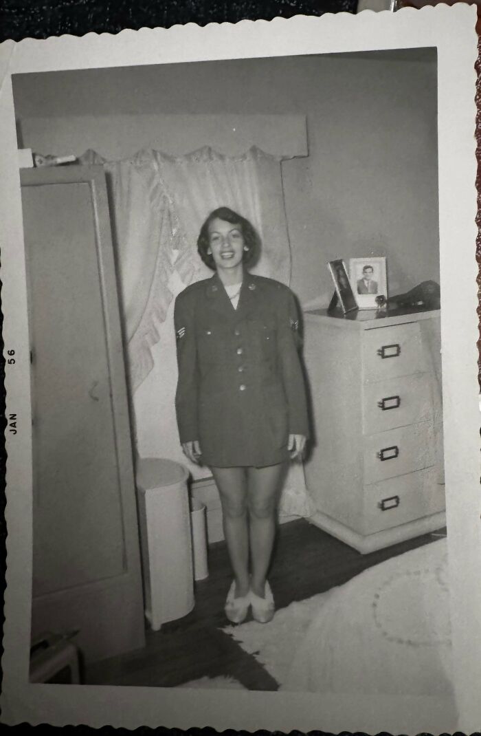 A historical photo of a woman in an oversized military jacket in a bedroom, smiling.
