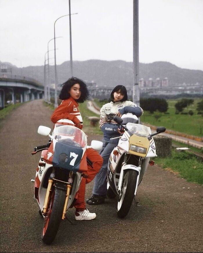 Two women pose with their motorcycles on a road, a compelling historical photo capturing a moment.
