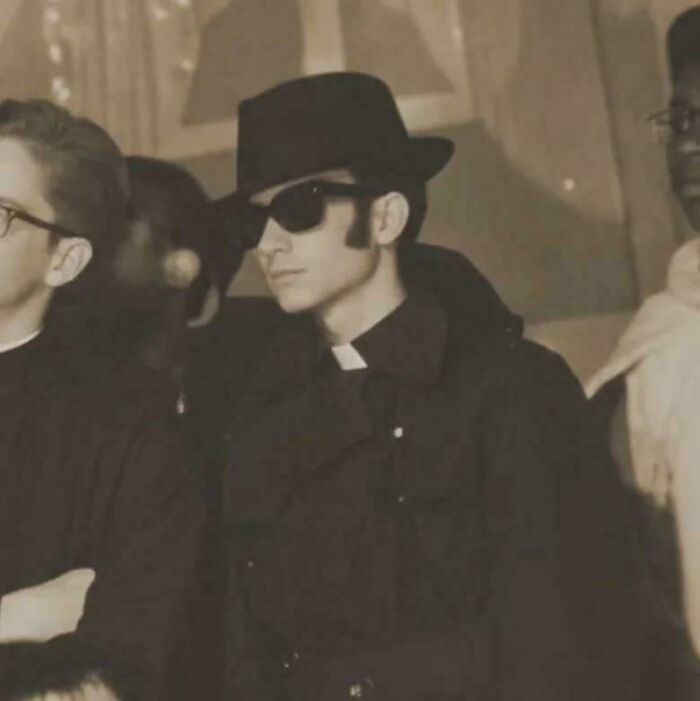A historical photo of a man in a black fedora, sunglasses, and a priest's collar, looking cool.