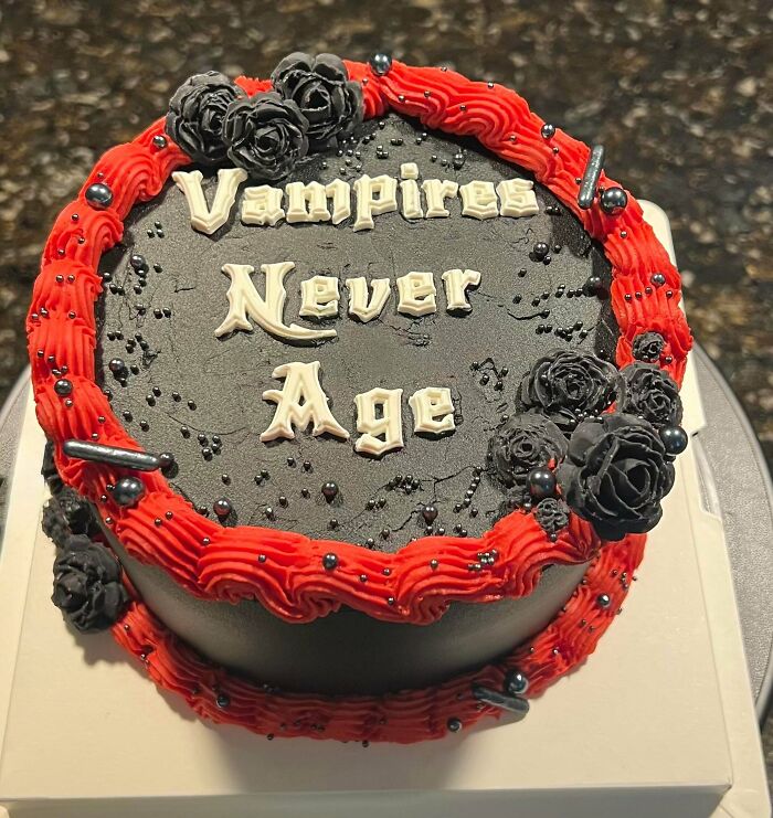 A gothic-themed cake with black icing, red trim, and the phrase 'Vampires Never Age,' showcasing amazing treats from bakers.
