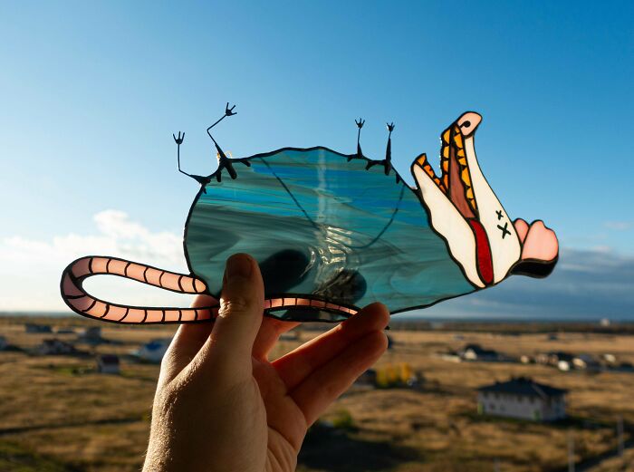 A person holds a stained-glass handmade project of a d**d rat against a clear blue sky, showing artistic talent and magic.