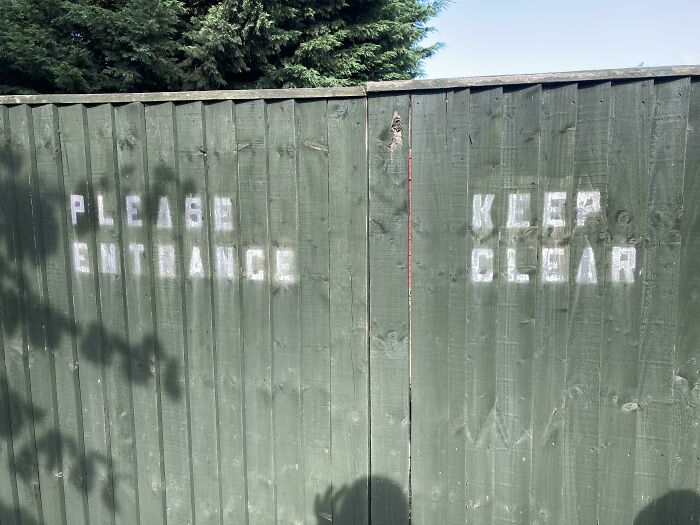 Faded stencil signage on green wooden fence reads please entrance keep clear
