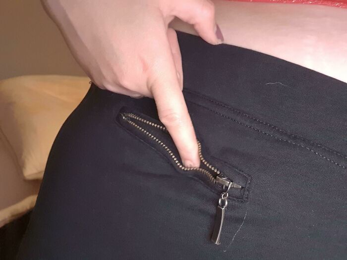 A close-up of a hand unzipping a tiny, non-functional zipper on black pants, showcasing bad clothing designs.