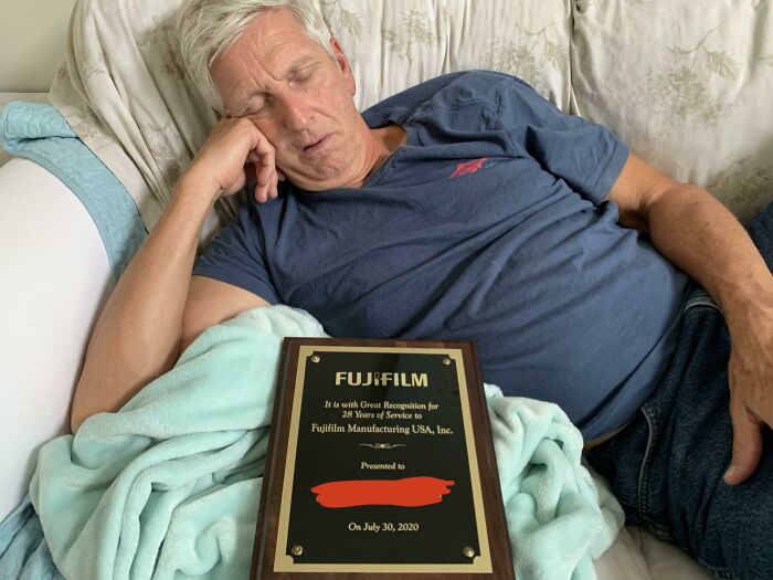 An amazing father naps on a couch with a Fujifilm award for 28 years of service on his lap.