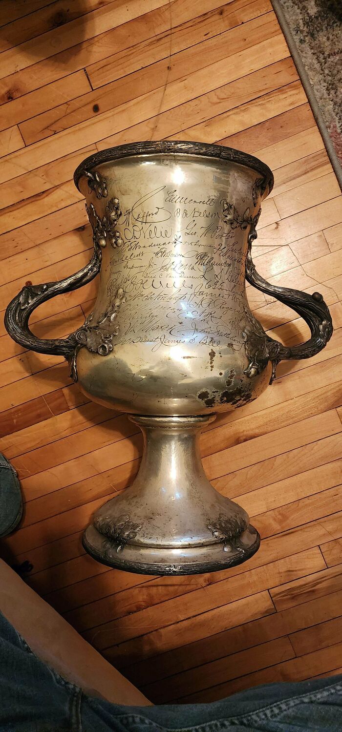 A large engraved silver trophy cup with ornate handles displayed on a wooden floor, appealing to antique lovers.