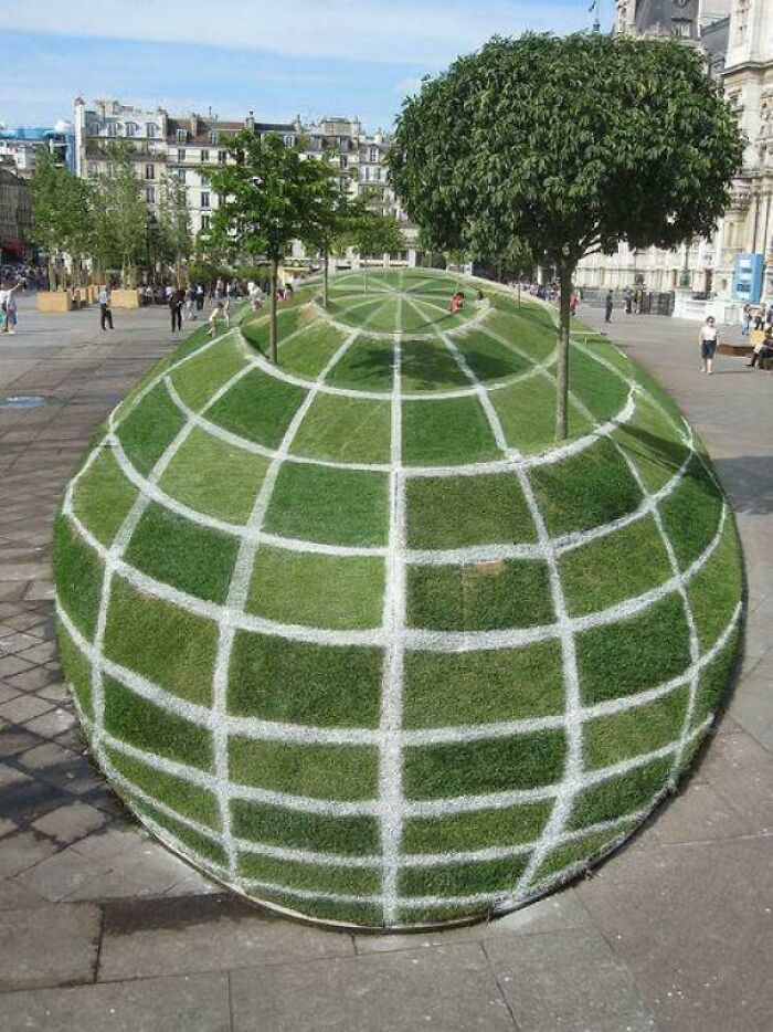 A large, grassy globe sculpture with white grid lines and trees, resembling a glitch in the matrix.