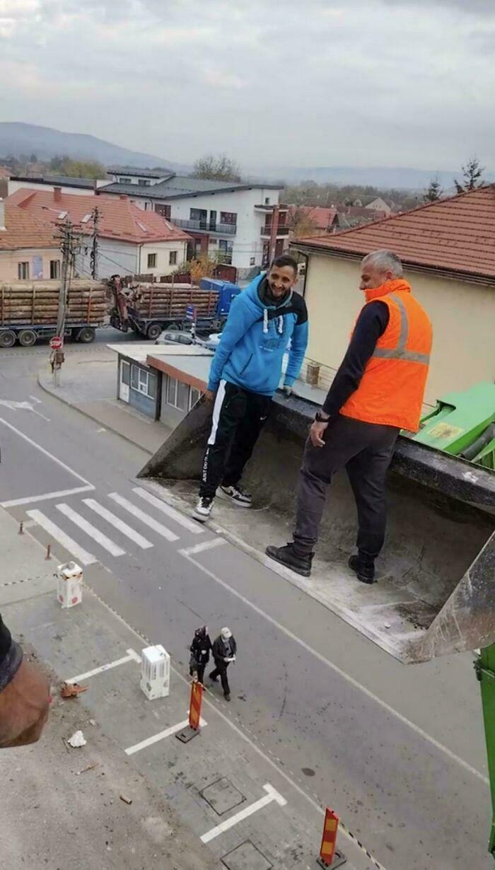 Two men stand precariously on a large bucket high above a street, an optical illusion or glitch in the matrix.