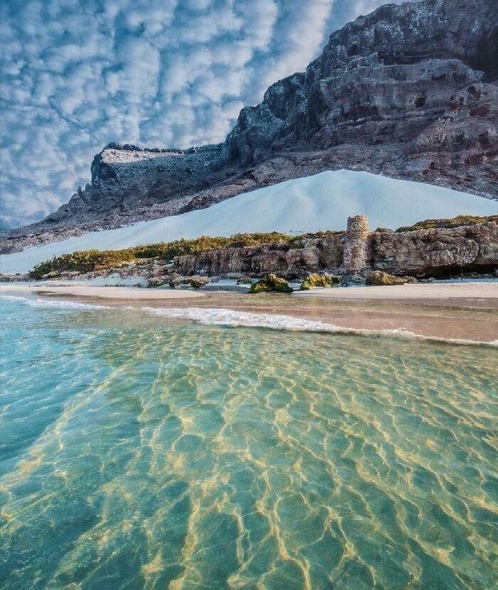 A stunning beach with clear water, white sand, and mountains in the background, resembling a Glitch in the Matrix.