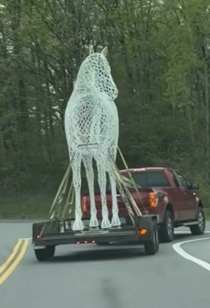 A large white wireframe horse sculpture on a trailer behind a red truck, resembling a glitch in the Matrix.