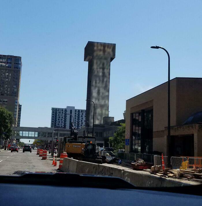 A tall, dark building in a city skyline resembling a giant T, creating a glitch in the matrix architectural illusion.