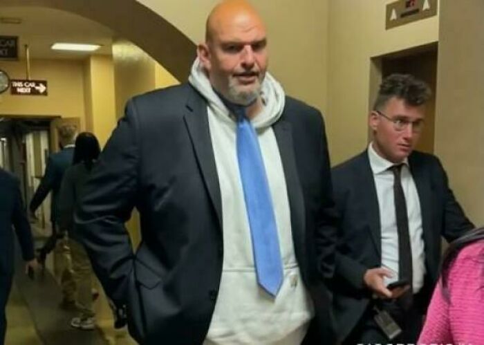 A peculiar man in a suit, hoodie, and light blue tie walking in a hallway, a real-life observation.