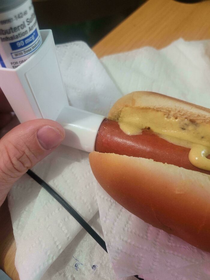 A hand holding an inhaler next to a hot dog with mustard, a peculiar photo from the “Not Interesting” community.