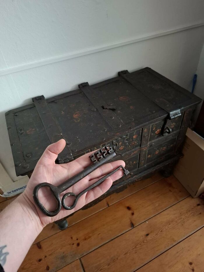 A hand holding large, antique iron keys in front of a rustic wooden chest filled with old treasures.