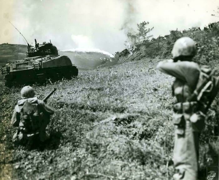 Two soldiers observe a tank firing a flamethrower on a hill during a battle, one of many historical photos.