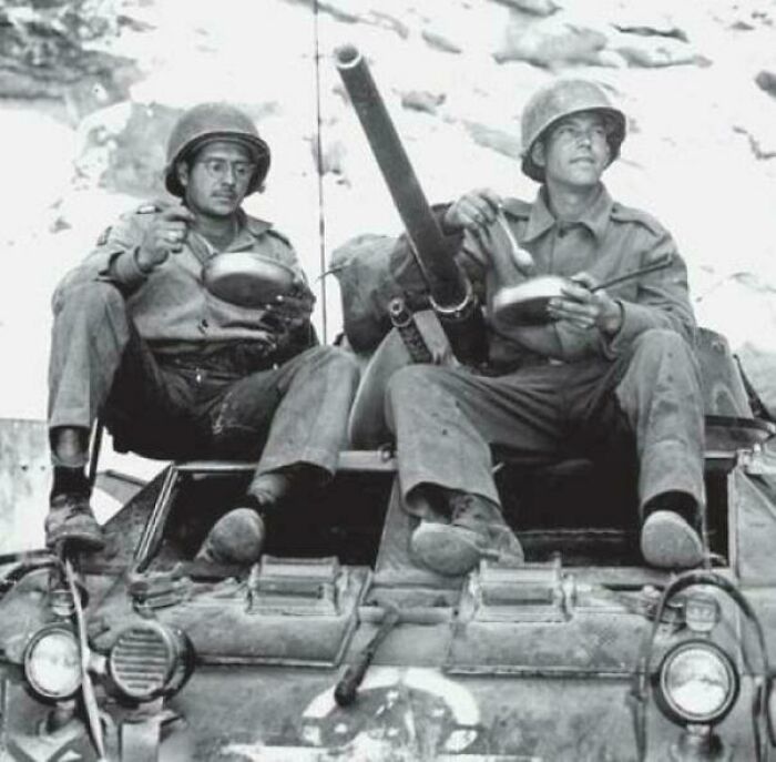 Two soldiers eat from bowls while sitting on a tank, a fascinating historical photo.