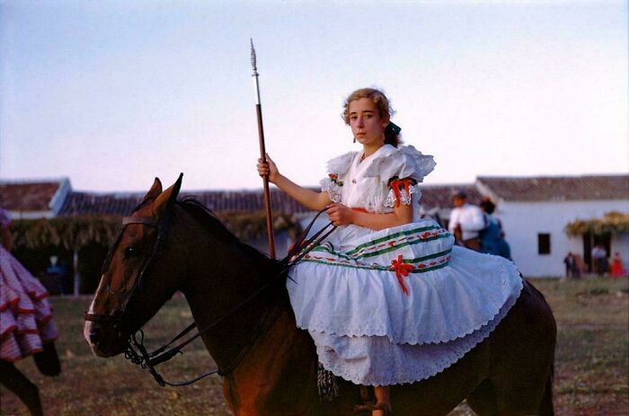 A girl in a white dress, holding a spear, rides a horse in an emotional historical photo.