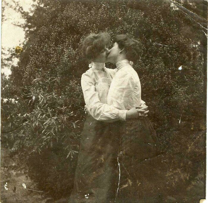 A black and white historical photo of two women embracing and kissing in front of lush foliage, a fascinating and emotional moment.