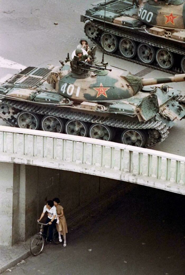 An emotional historical photo shows two military tanks driving on a road while people on bicycles pass under a bridge below.
