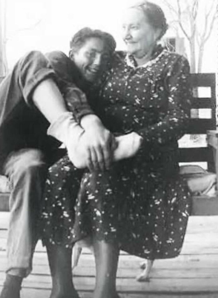 A black and white historical photo shows a young man smiling while being held by an older woman sitting on a bench.