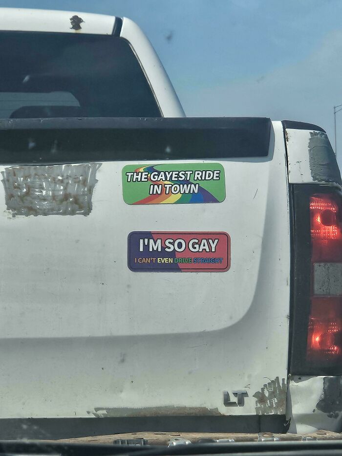 The back of a white pickup truck showing two unfiltered bumper stickers about being gay.
