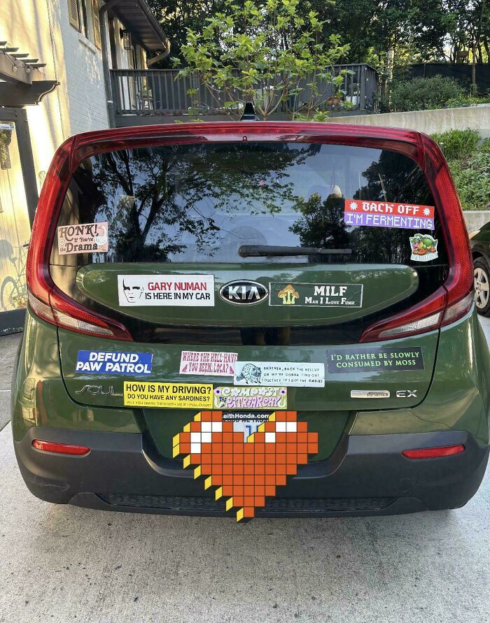 A green Kia Soul car with many wild bumper stickers covering its rear window and bumper.
