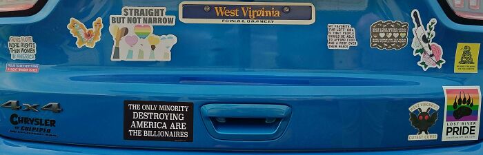 A blue car's bumper displays several funny bumper stickers, including political and pride-themed ones.