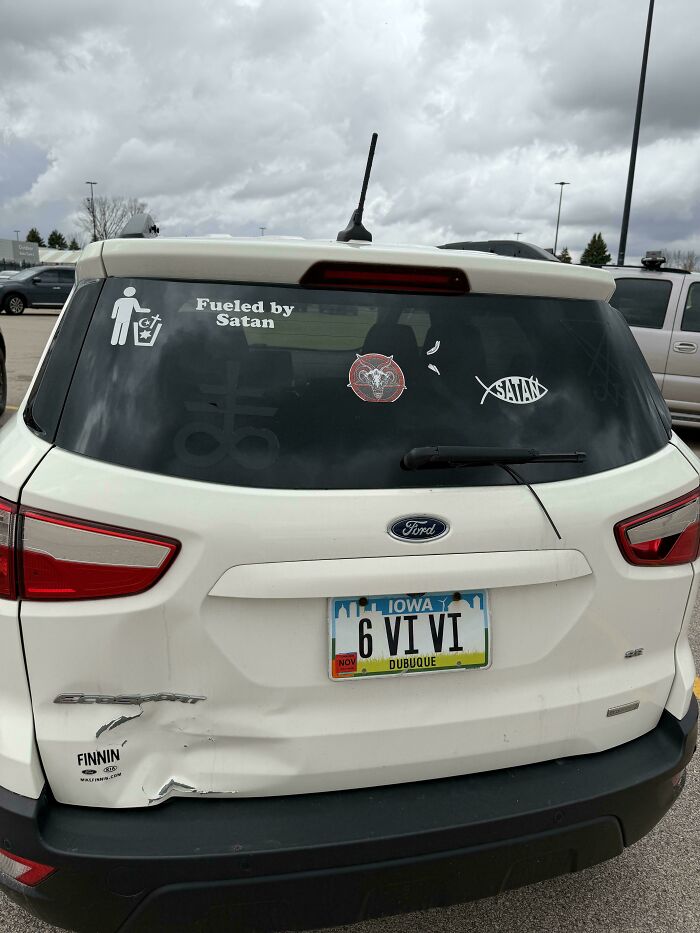 Unfiltered bumper stickers on a white Ford, including Fueled by Satan and 6 VI VI on the license plate.