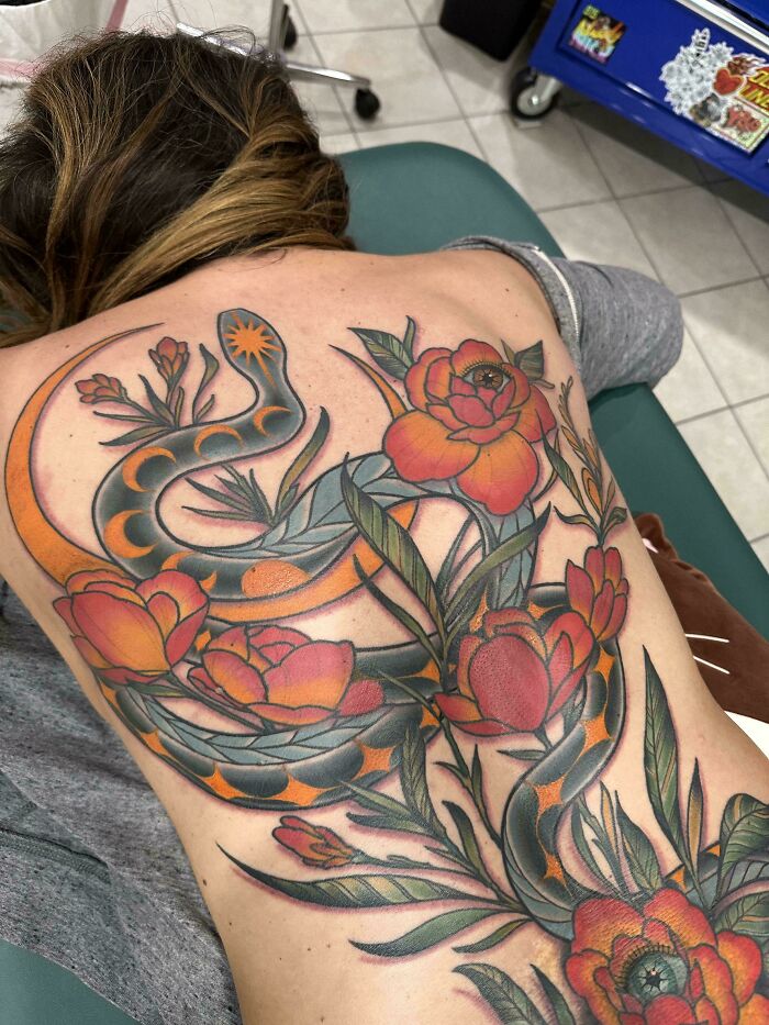 A detailed back piece tattoos featuring a snake intertwined with large, colorful flowers on a person's back. Stunning tattoo.