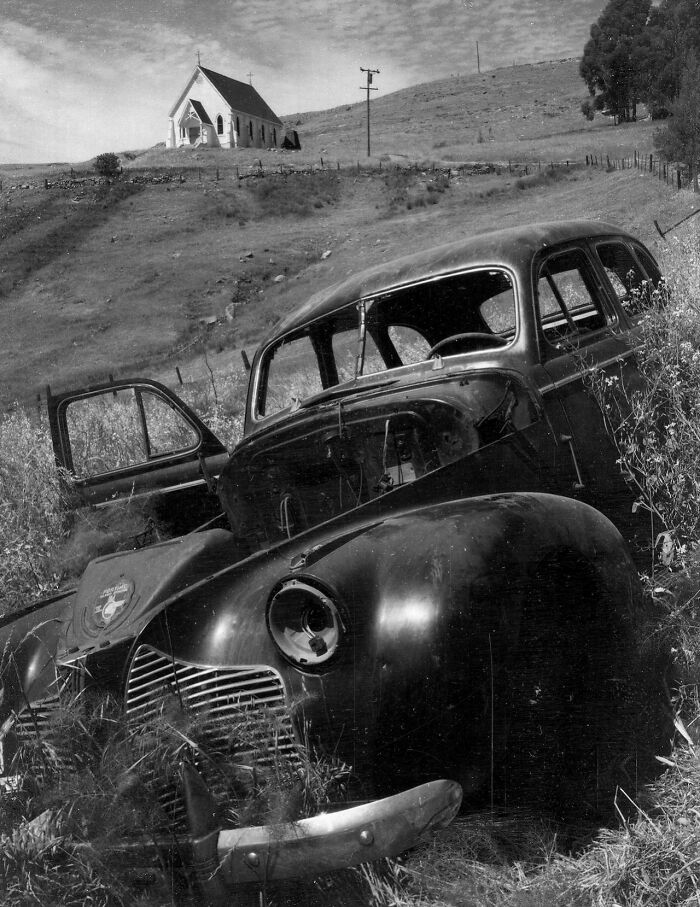 Old rusty car abandoned in field near church in vintage photo