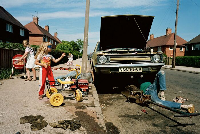 Children playing near broken car with open hood on residential street