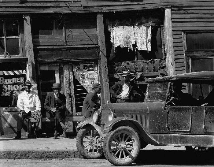 Vintage photo showing men sitting by old car in front of barber shop