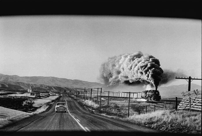 Black and white photo of a train and car on a rural road with vast landscape