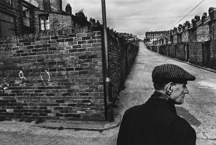 Black and white image of man in flat cap walking past brick walls in old neighborhood