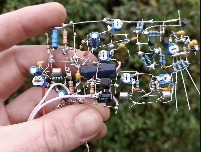 A person holding a circuit board with messy wiring and components, illustrating a handmade electronics fail.