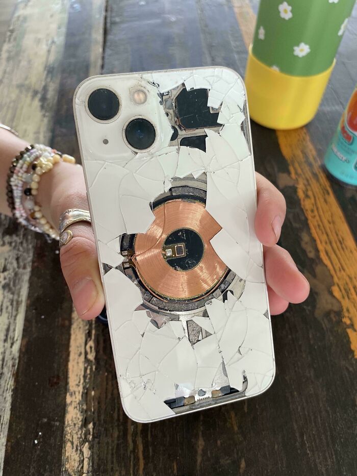A hand holding a white iPhone with a severely cracked back, revealing the internal components, illustrating Electronics Fails.