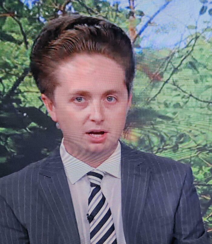 A man with a very tall hairstyle, appearing to be a hair salon disaster, wearing a suit and striped tie.
