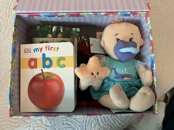 A baby doll, ABC book with an apple, and star toy in a decorative box, symbolizing the start of new families from adoption stories.
