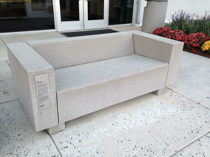 A solid concrete couch with a plaque, positioned outdoors near a building entrance. An interior designer might appreciate its minimalist form.