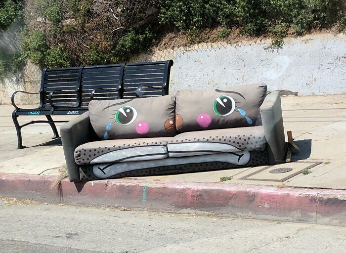 A discarded couch with painted crying faces sits by a curb, next to a black metal bench. An interior designer might see potential in its art.